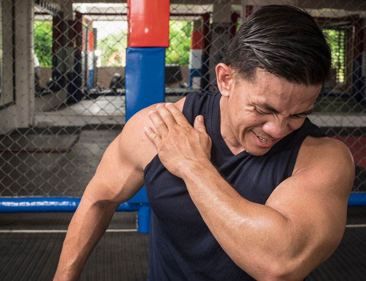An athletic man in a gym clutches his shoulder with a pained expression, illustrating a workout injury like a rotator cuff tear.